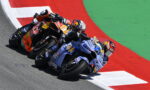 Three professional motorcycle racers lean into a tight turn on a race track, orange, blue, and red bikes close together.