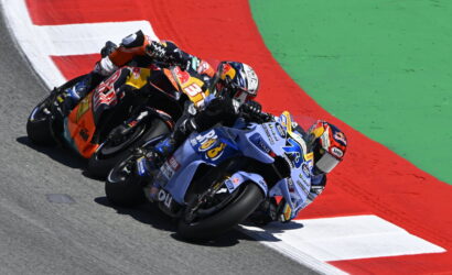 Three professional motorcycle racers lean into a tight turn on a race track, orange, blue, and red bikes close together.