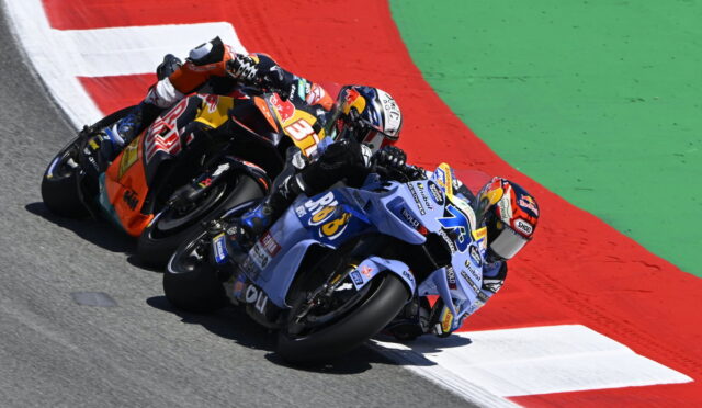 Three professional motorcycle racers lean into a tight turn on a race track, orange, blue, and red bikes close together.