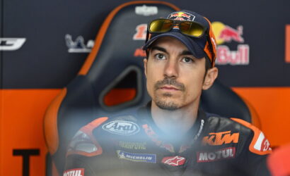 MotoGP rider in a KTM racing suit with Red Bull logos, seated in a chair against a branded backdrop.