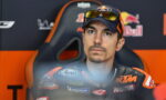 MotoGP rider in a KTM racing suit sits in a team chair, wearing a cap and sunglasses, looking upward with a focused expression.