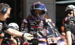 MotoGP rider in a black-and-purple racing suit and helmet sits on a race bike in the pit, a crew member reaches toward the bike beside him.