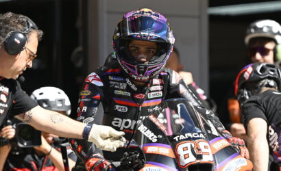 MotoGP rider in a black-and-purple racing suit and helmet sits on a race bike in the pit, a crew member reaches toward the bike beside him.