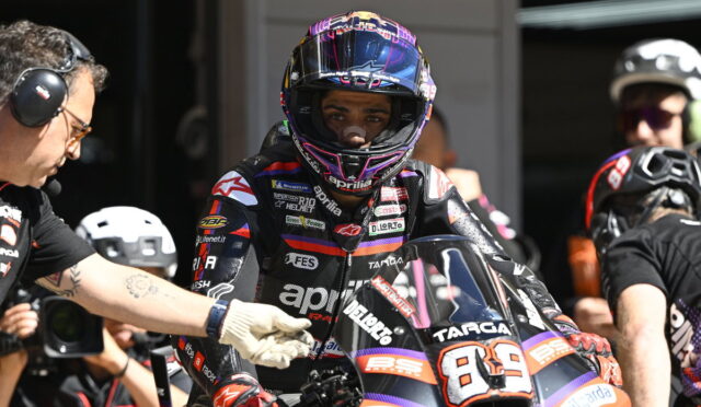 MotoGP rider in a black-and-purple racing suit and helmet sits on a race bike in the pit, a crew member reaches toward the bike beside him.