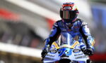 Moto racer in blue racing suit on a blue bike, number 13, wearing a red‑white Shoei helmet with sponsor logos.