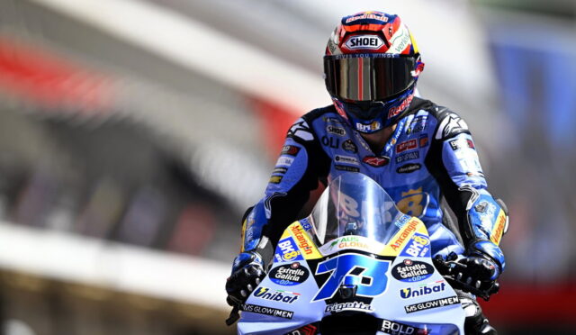 Motorcycle racer in blue racing suit and full-face helmet on a blue-and-white race bike, number 13, on track.