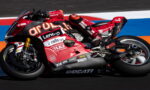 MotoGP rider leaning into a red Ducati racing bike on a colorful race track with sponsor logos visible on the bike's fairing