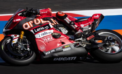MotoGP rider leaning into a red Ducati racing bike on a colorful race track with sponsor logos visible on the bike's fairing