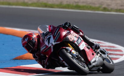 Racer in red suit leans into a sharp turn on a red Ducati motorcycle at a race track, blue and orange curbs visible nearby.
