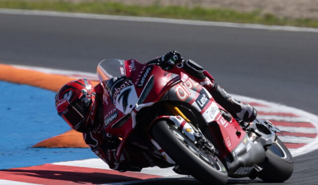 Racer in red suit leans into a sharp turn on a red Ducati motorcycle at a race track, blue and orange curbs visible nearby.