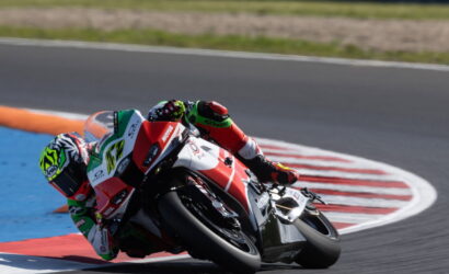 Motorcycle racer in green, red, and white leans into a sharp turn on a race track, knee nearly touching the asphalt.