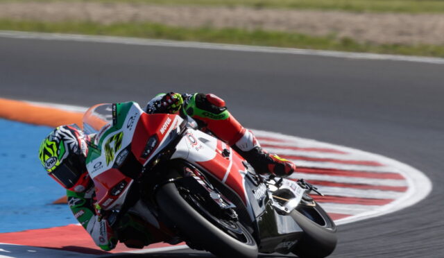 Motorcycle racer in green, red, and white leans into a sharp turn on a race track, knee nearly touching the asphalt.