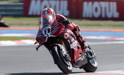 Motorcycle racer in red leathers leaning into a track corner on a red race bike, number 77 visible.