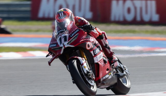 Motorcycle racer in red leathers leaning into a track corner on a red race bike, number 77 visible.