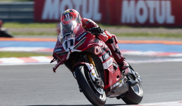 Racer on a red racing motorcycle leaning into a turn on a track, with sponsor logos visible on the bike's fairings