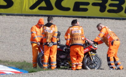 Five racers' crew in bright orange overalls work on a red sport motorcycle on a gravel pit area by a large yellow banner.