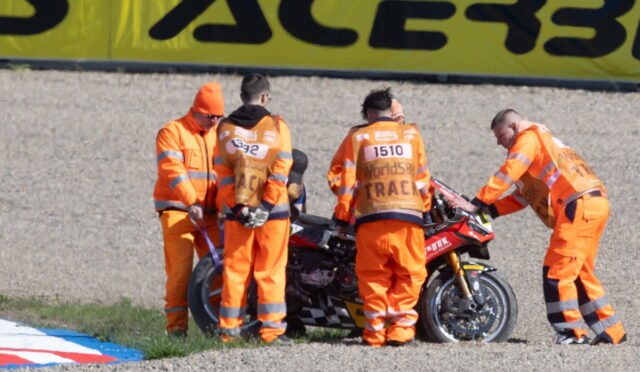 Five racers' crew in bright orange overalls work on a red sport motorcycle on a gravel pit area by a large yellow banner.