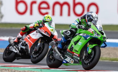 Two motorcycle racers lean into a curve on a race track, green bike in front with sponsor decals, red-and-white bike close behind.