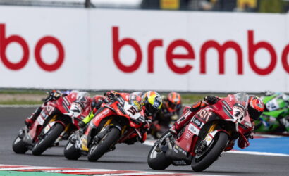 Motorcycle race: four riders lean into a turn on a track, red bikes with sponsor logos and Brembo signage in the background.