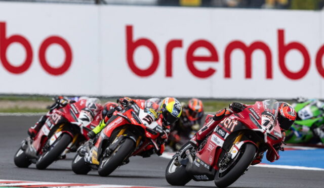 Motorcycle race: four riders lean into a turn on a track, red bikes with sponsor logos and Brembo signage in the background.