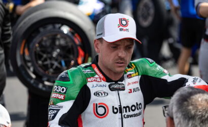 Motorcycle racer in green-white Bimota Kawasaki suit, wearing a white cap, standing in pit area with a wheel in the background.