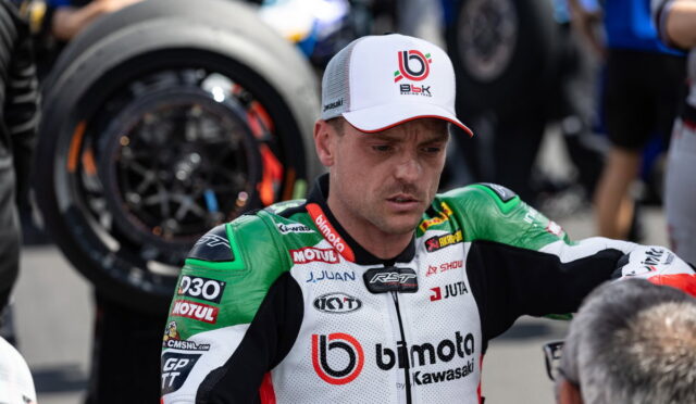 Motorcycle racer in green-white Bimota Kawasaki suit, wearing a white cap, standing in pit area with a wheel in the background.