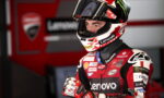 Moto racer in red and black racing suit with Lenovo branding, wearing a helmet, standing in a pit area.