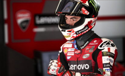 Moto racer in red and black racing suit with Lenovo branding, wearing a helmet, standing in a pit area.