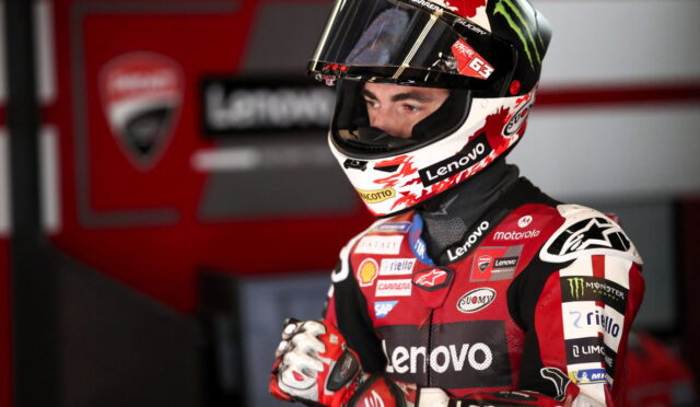 Moto racer in red and black racing suit with Lenovo branding, wearing a helmet, standing in a pit area.