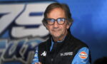 Portrait of a man in a blue racing team jacket and glasses, standing against a blue backdrop.