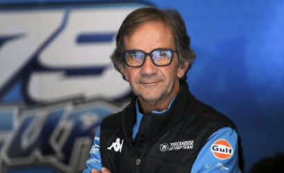 Portrait of a man in a blue racing team jacket and glasses, standing against a blue backdrop.