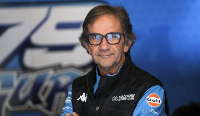 Portrait of a man in a blue racing team jacket and glasses, standing against a blue backdrop.