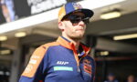 Racing driver wearing a blue and orange KTM/Red Bull team polo, cap, and dark sunglasses, standing in a pit area.