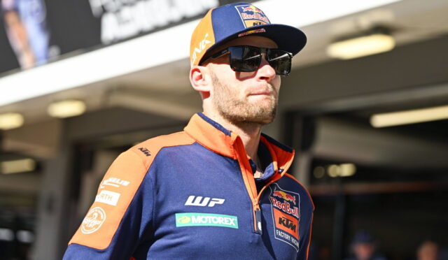 Racing driver wearing a blue and orange KTM/Red Bull team polo, cap, and dark sunglasses, standing in a pit area.
