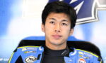 Young Asian motorcycle racer in a blue suit with sponsor logos, looking upward, against a blue backdrop.