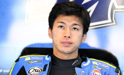 Young Asian motorcycle racer in a blue suit with sponsor logos, looking upward, against a blue backdrop.
