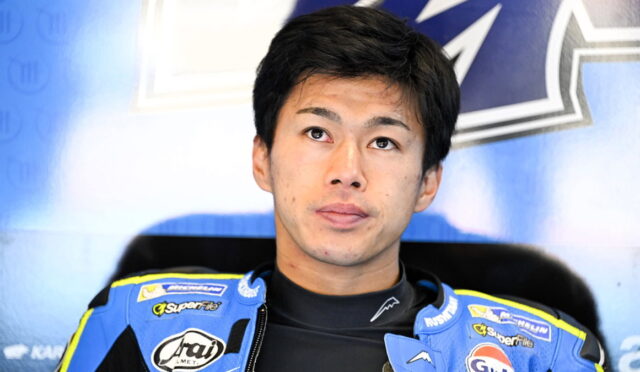 Young Asian motorcycle racer in a blue suit with sponsor logos, looking upward, against a blue backdrop.