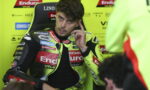 Racer in a neon-yellow racing suit sits in a paddock chair, touching his ear as he talks.