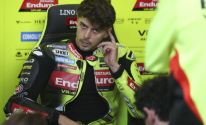Racer in a neon-yellow racing suit sits in a paddock chair, touching his ear as he talks.