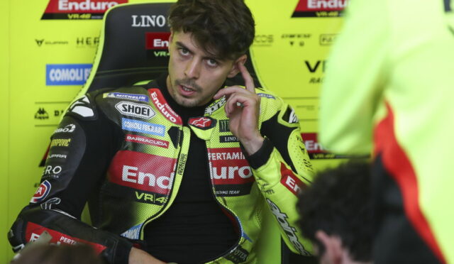 Racer in a neon-yellow racing suit sits in a paddock chair, touching his ear as he talks.