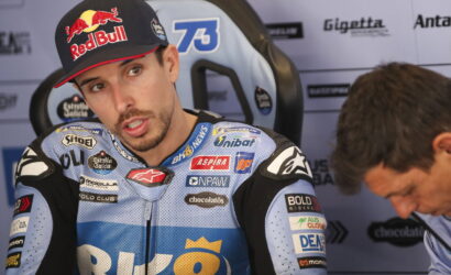 Racer wearing a blue racing suit with sponsor logos and a Red Bull cap, seated in a interview chair outdoors/garage area.