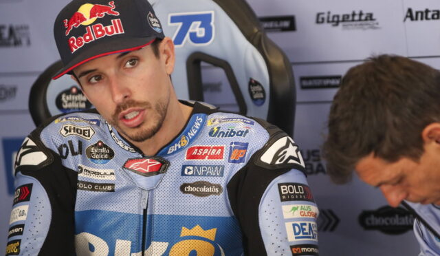 Racer wearing a blue racing suit with sponsor logos and a Red Bull cap, seated in a interview chair outdoors/garage area.