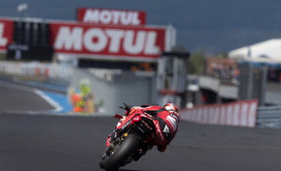 Motorcycle racer in a red suit leaning into a turn on a race track with Motul banners in the background.