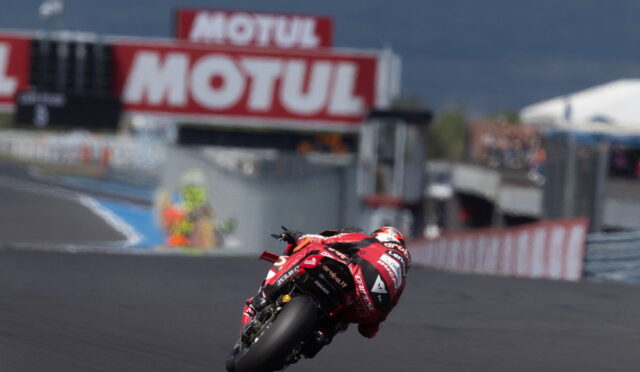Motorcycle racer in a red suit leaning into a turn on a race track with Motul banners in the background.