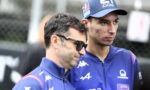 Two men in blue racing team jackets stand side by side outdoors, one wearing sunglasses and a cap with sponsor logos.