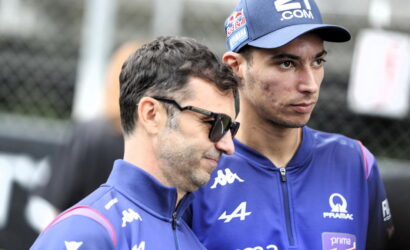 Two men in blue racing team jackets stand side by side outdoors, one wearing sunglasses and a cap with sponsor logos.