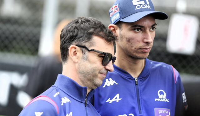 Two men in blue racing team jackets stand side by side outdoors, one wearing sunglasses and a cap with sponsor logos.