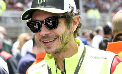 Close-up of a man in a neon-yellow racing jacket and cap, smiling in sunglasses at a crowded outdoor event.