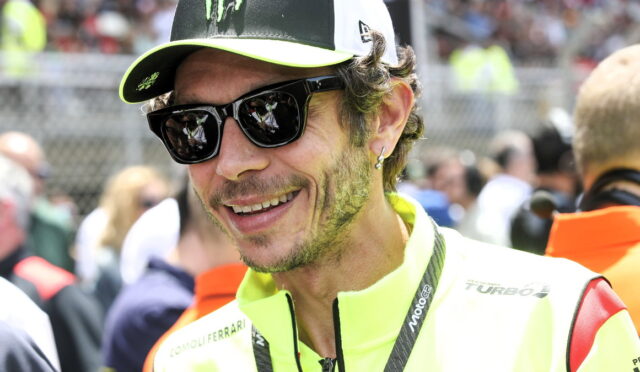 Close-up of a man in a neon-yellow racing jacket and cap, smiling in sunglasses at a crowded outdoor event.