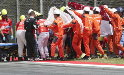 Pit crew in orange suits and medical staff carry a white sheet over a stretcher beside a crash on the track.